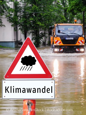 Ein Warnschild mit der Aufschrift 'Klimawandel' vor einer überfluteten Straße, auf der ein Einsatzfahrzeug durch das Wasser fährt.