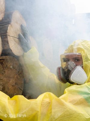 Person in Schutzanzug und Maske steht vor einem Container mit Holz, aus dem Rauch entweicht.