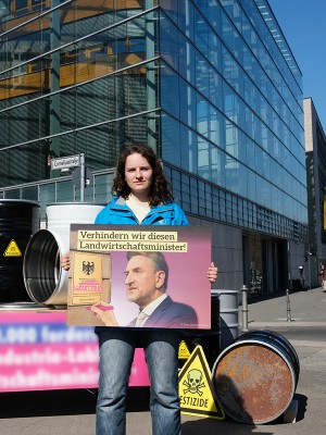 Person steht mit einem Schild in der Hand vor einem Gebäude. Auf dem Schild steht "Verhindern wir diesen Agrarminister!"