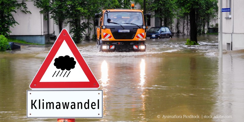 Ein Warnschild mit der Aufschrift 'Klimawandel' vor einer überfluteten Straße, auf der ein Einsatzfahrzeug durch das Wasser fährt.