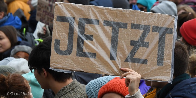 Bild zeigt eine Demonstration, im Vordergrund wird ein Schild mit "JETZT" hochgehalten