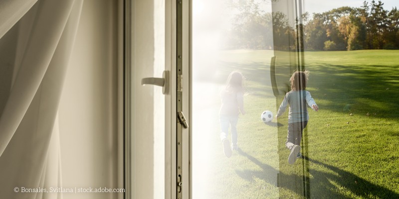 Blick durch ein geöffnetes Fenster: Zwei Kinder spielen draußen auf einer sonnigen Wiese mit einem Ball.
