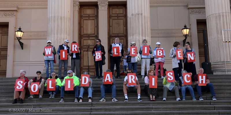 Demonstrierende sitzen und stehen auf den Stufen vor einem öffentlichen Gebäude und halten rote Buchstabentafeln, die zusammen den Satz „Wir bleiben solidarisch“ ergeben.